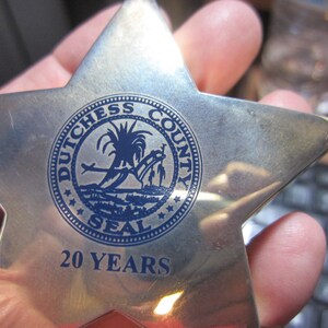 Puede incluir: Una insignia plateada en forma de estrella con el Sello del Condado de Dutchess en azul. El sello presenta un diseño de palmera y puente, con el texto "20 YEARS" en la parte inferior. La insignia se sostiene en una mano.