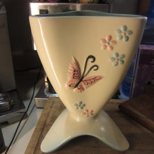 May include: A white ceramic vase with a triangular shape and a blue and pink butterfly design. The vase has a light blue glaze and is decorated with pink and blue flowers.