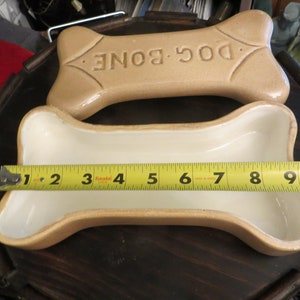 May include: A ceramic dog bone shaped trinket box with a lid. The lid is beige and has the words "DOG BONE" embossed on it. The box is beige and white.