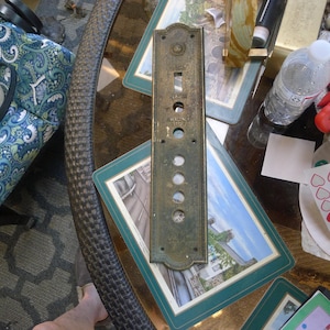 May include: A rectangular, weathered brass elevator control panel featuring a series of circular cutouts and vertical slots. The metal plate includes engraved text labels reading LIGHT, ALARM, and EMERGENCY STOP, with small screw holes at the top and bottom.