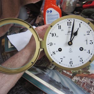 May include: A round, brass-colored clock with a white face and black numerals. The clock has a hinged, circular glass cover. The clock face displays the text "Royal Marine" and the time is approximately 1:00. The clock is a vintage nautical item.