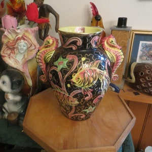 May include: A black ceramic vase with a yellow, pink, and green floral design. The vase has two seahorse handles and a yellow rim. The vase is sitting on a wooden table.
