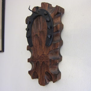 May include: A wooden wall decor with a horseshoe attached to it. The horseshoe is black metal with nail heads. The wood is a dark brown color.