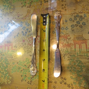 May include: Two silver butter knives with ornate handles, one with a plain blade and the other with a decorative blade, are shown next to a yellow measuring tape.