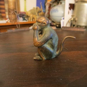 May include: A small bronze monkey figurine with a green patina. The monkey is sitting with its hands clasped in front of its face, and its tail is curled around its body.