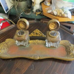 May include: A vintage brass inkwell set with two glass inkwells and a decorative tray. The inkwells have lids with ornate designs and the tray has a scalloped edge.