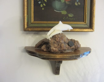 May include: A white dolphin sculpture leaping from a textured, brown wooden base. The sculpture sits on a small, ornate wooden shelf. A framed painting with a gold border hangs above the shelf.