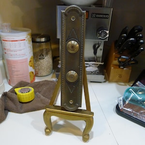 May include: An antique brass door plate with two round push buttons is displayed on a small, gold-painted easel. The door plate has a rectangular shape with decorative trim and a vintage aesthetic. The easel has curved legs and a simple design.