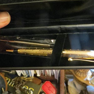 May include: A gold-toned ballpoint pen with a textured, grooved barrel and a black tip, presented in a black rectangular case. The pen features a gold clip and is accompanied by a spare ink refill encased in a clear plastic sleeve.