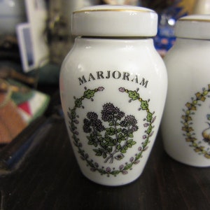 May include: Two white ceramic spice jars with gold lids and floral designs. The jar on the left says "MARJORAM" and the jar on the right says "CHIVE".