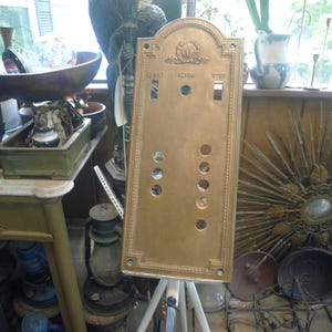 May include: A rectangular brass elevator control panel featuring an arched top with an embossed laurel wreath emblem. The plate includes the engraved words LIGHT, ALARM, and STOP above rectangular and circular cutouts for buttons and switches.