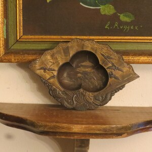 May include: A small, ornate, bronze-coloured trinket dish with a fan-shaped design. The dish has three compartments, each with a different design. The dish is sitting on a wooden shelf. The text "L. Rygjek" is visible in the background.