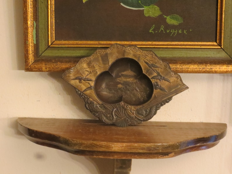 May include: A small, ornate, bronze-coloured trinket dish with a fan-shaped design. The dish has three compartments, each with a different design. The dish is sitting on a wooden shelf. The text "L. Rygjek" is visible in the background.