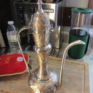 May include: A silver and gold ornate teapot with a floral design. The teapot has a long, curved spout and a lid with a finial.