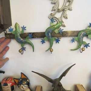 May include: Three green and blue metal gecko lizard hooks mounted on a brown metal bar. The hooks are shaped like geckos with their tails curled up. The geckos have gold accents and are attached to the bar with screws.