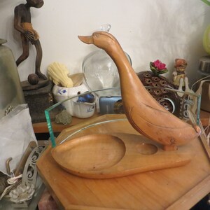 May include: A wooden duck figurine with a carved design. The duck is holding a small wooden dish with a round indentation. The duck is sitting on a wooden base.