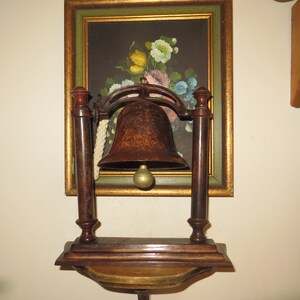 May include: A decorative bell with a dark brown finish and a gold-colored clapper, mounted on a wooden stand with a gold trim. The bell is hanging from a white rope.