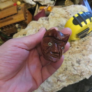 May include: A hand holds a small wooden carving of a face with exaggerated features. The carving has a brown color and white eyes.