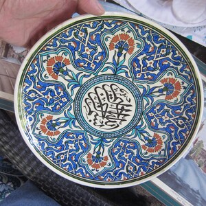 May include: Decorative ceramic plate with a detailed floral and geometric pattern in blue, orange, and white. The plate features a central design with black calligraphy. The rim is green.