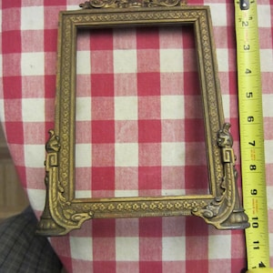 May include: A gold-toned ornate picture frame with a decorative design. The frame is on a red and white checkered tablecloth.