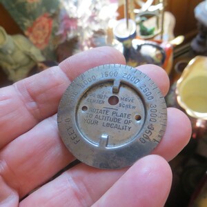 May include: A round metal plate with a dial marked in feet from 0 to 3500. The dial has a center hole and two smaller holes. The text on the dial reads "DO NOT CENTER", "MOVE SCREW", "ROTATE PLATE TO ALTITUDE OF YOUR LOCALITY".