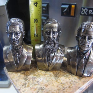 May include: Three silver-toned bust sculptures of men, each with a different hairstyle and facial features. The sculptures are displayed on a light brown surface.