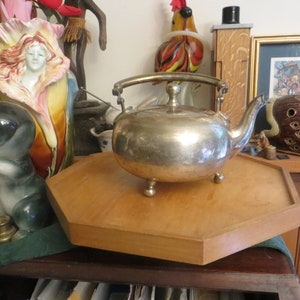 May include: A silver teapot with a wooden octagonal base. The teapot has a round body with a curved handle and a spout. The teapot is sitting on a wooden table with other decorative items.