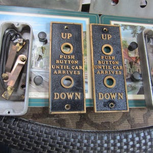 May include: Vintage elevator call buttons with "UP" and "DOWN" text. The buttons are black with gold lettering and trim. Two metal boxes with electrical components are also visible, along with screws and other hardware.