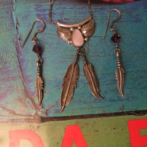 May include: A silver-toned jewelry set featuring a necklace and matching earrings. The necklace has a central pendant with a light pink stone and two feather charms. The earrings each have a dangling feather charm and a dark red crystal.