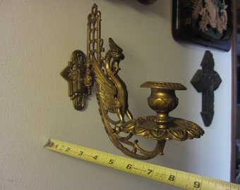 May include: A gold-toned metal wall sconce with a bird design and a candle holder. The sconce is attached to a wall with a screw and has a decorative design.