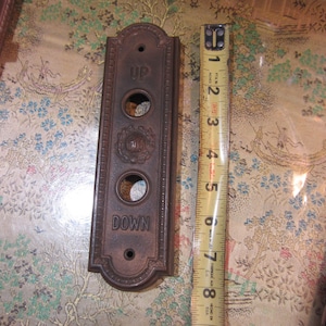 May include: A vintage bronze elevator button panel with the words "UP" and "DOWN" and a button for the first floor. The panel is approximately 8 inches long. A yellow measuring tape is next to the panel.