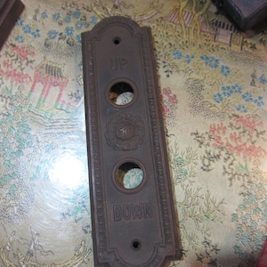 May include: A rectangular, antique elevator button panel made of dark metal. The panel has three circular button holes and the words "UP" and "DOWN" are engraved. A decorative emblem is centered between the buttons.