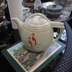 May include: A white ceramic teapot with a lid and a handle. The teapot has a design of two red birds on a branch. The teapot is sitting on a green coaster with a picture of a house.