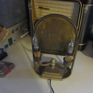 May include: Vintage brass wall sconce with two light bulbs. The sconce has a curved backplate and a decorative base. The bulbs are teardrop-shaped. The sconce is designed to be hardwired and has a black cord.