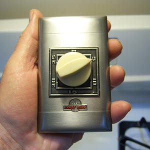 May include: A silver-colored kitchen timer with a cream-colored dial and a black and silver face. The timer has a rectangular shape and the brand name "Trade Wind" is visible. The timer is held in a hand.