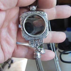 Pu&ograve; includere: Una collana con pendente argentato con cornice quadrata e specchio rotondo. La cornice presenta volute ornate e un nappa decorativa. Il pendente &egrave; sospeso a una catena delicata, creando un accessorio elegante.