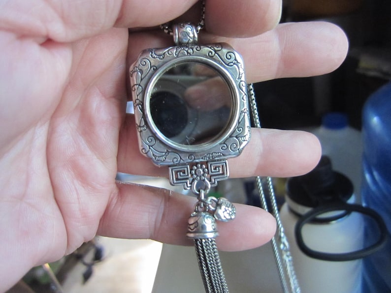 Pu&ograve; includere: Una collana con pendente argentato con cornice quadrata e specchio rotondo. La cornice presenta volute ornate e un nappa decorativa. Il pendente &egrave; sospeso a una catena delicata, creando un accessorio elegante.