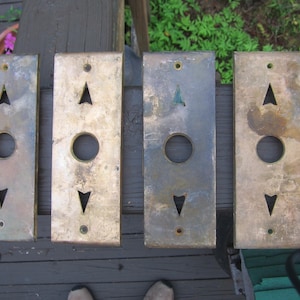 May include: Four vintage elevator call plates with a rectangular shape. Each plate is made of tarnished brass and features an arrow pointing up, a circular button, and an arrow pointing down.
