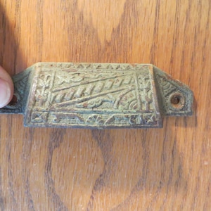 May include: A green metal drawer pull with an ornate design. The pull has a rectangular shape with rounded edges and a decorative pattern on the front.