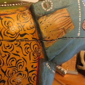 Puede incluir: Una figurita de elefante de madera pintada en azul, amarillo y naranja. El elefante tiene un patr&oacute;n floral en su cuerpo y una grieta en el medio.