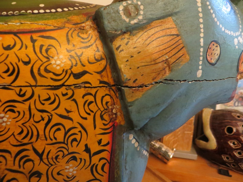 Puede incluir: Una figurita de elefante de madera pintada en azul, amarillo y naranja. El elefante tiene un patr&oacute;n floral en su cuerpo y una grieta en el medio.