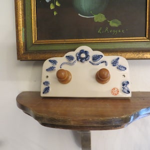 May include: White ceramic wall-mounted coat rack with blue floral designs and two wooden knobs. The rack is mounted on a wooden shelf.