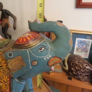 Puede incluir: Figurita de elefante de madera pintada a mano en colores azul, verde, amarillo, naranja y rojo. El elefante tiene un dise&ntilde;o detallado con patrones intrincados y una guirnalda de cuentas alrededor del cuello.