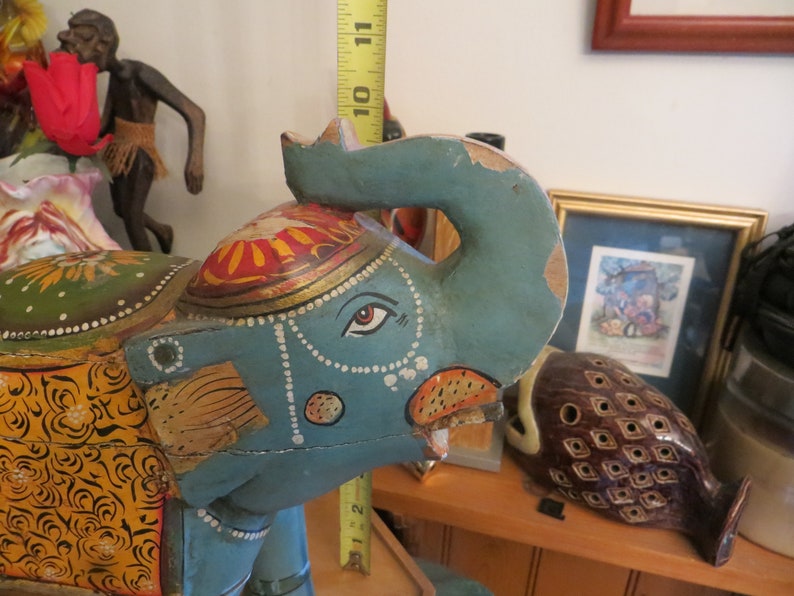 Puede incluir: Figurita de elefante de madera pintada a mano en colores azul, verde, amarillo, naranja y rojo. El elefante tiene un dise&ntilde;o detallado con patrones intrincados y una guirnalda de cuentas alrededor del cuello.