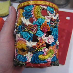 May include: A cylindrical ceramic container with a red base and a gold rim. The container is decorated with a colorful, intricate design of dragons and clouds.