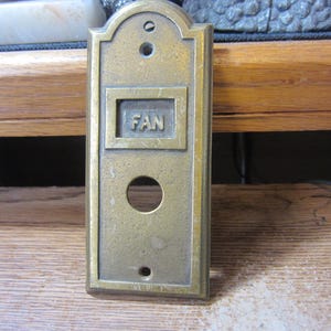 May include: Vintage brass fan switch plate with the word "FAN" in a recessed square. The plate has a rectangular shape with a rounded top and a single large circular hole. There are two smaller holes above and one below the circular hole.
