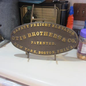 May include: An oval, bronze-colored sign with gold lettering, reading "SAFETY FREIGHT ELEVATOR OTIS BROTHERS & CO. PATENTED. NEW YORK, BOSTON, PHIL'A." The sign is mounted on a small metal stand.