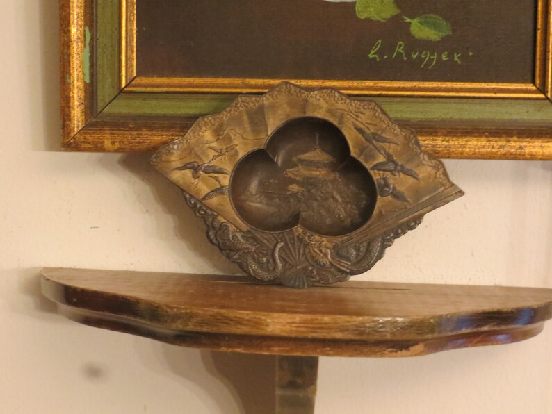 May include: A small, ornate, gold-coloured metal trinket dish shaped like a fan with three compartments. The dish features a detailed design of birds and flowers. The dish is sitting on a wooden shelf.
