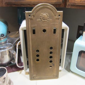 May include: A brass elevator control panel with a decorative design. The panel has buttons for "Light", "Alarm", and "Emergency Stop".