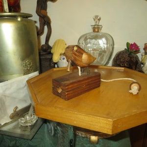 May include: A wooden bird toy with a pull string, perched on a wooden block with metal springs for legs. The bird is brown with a brass-colored eye. The toy sits on a wooden hexagonal tray.
